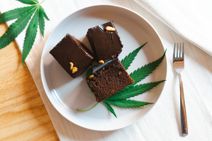 Chocolate brownies on a plate with cannabis leaves and a fork on a wooden surface.